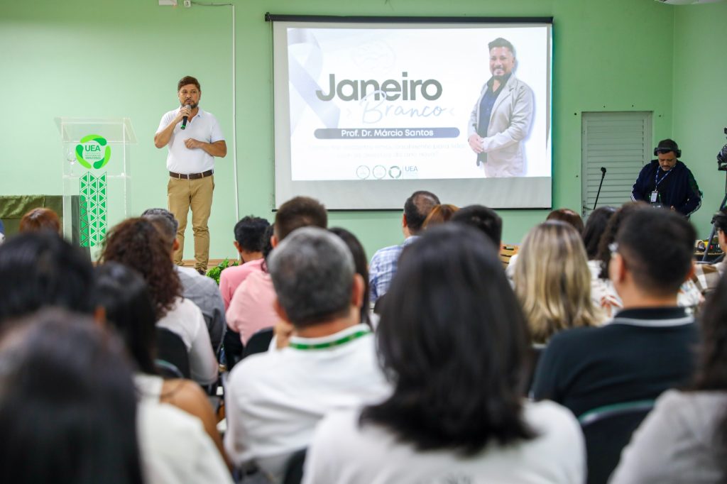 Fotografia em plano médio do auditório da Reitoria durante o Janeiro Branco. À esquerda, o palestrante, Prof. Dr. Márcio Gonçalves, em pé e segurando um microfone, fala para o público. Ao fundo, uma tela de projeção exibe o slide de abertura com o título "Janeiro Branco" e a foto do palestrante. Em primeiro plano, as costas e cabeças dos participantes sentados criam profundidade, focando a atenção na apresentação sobre bem-estar e saúde mental.