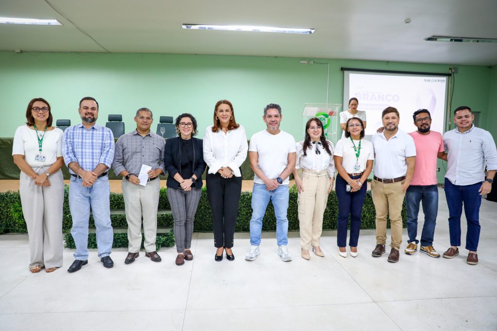 Fotografia de grupo em plano médio e horizontal, tirada num auditório com parede verde ao fundo. No centro da parede, uma tela de projeção exibe o logótipo da campanha "Janeiro Branco". Onze servidores da UEA, homens e mulheres, estão alinhados lado a lado, sorridentes. A maioria veste camisas brancas e calças de ganga escuras, utilizando crachás de identificação pendurados no pescoço. O ambiente é iluminado e sugere o encerramento da palestra sobre saúde mental realizada na Reitoria.