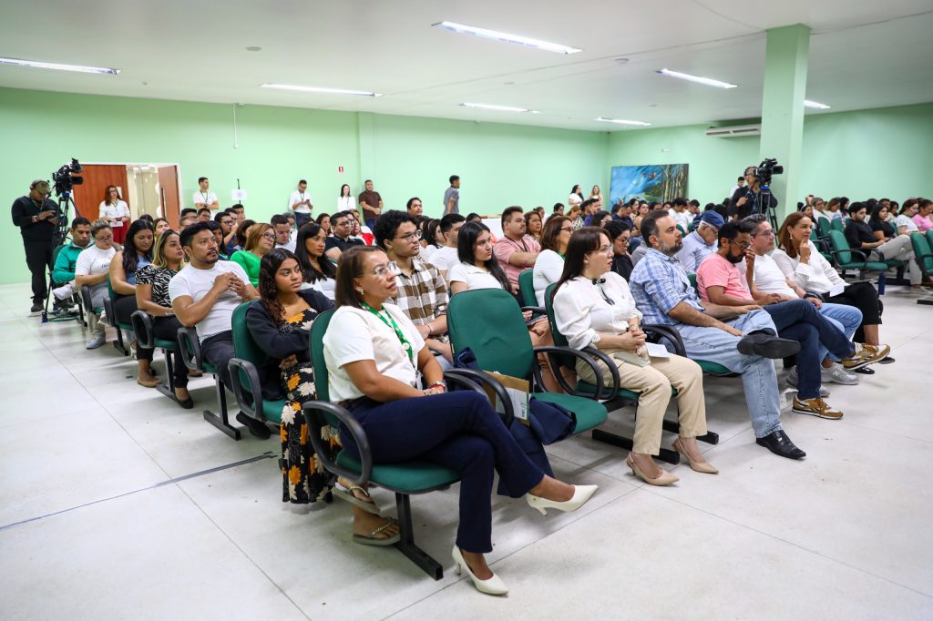 Fotografia de plano aberto do auditório da Reitoria da UEA lotado durante a palestra do Janeiro Branco. Dezenas de servidores e membros da comunidade acadêmica estão sentados em poltronas verdes, acompanhando atentamente a programação voltada ao bem-estar e saúde mental. No lado esquerdo, um cinegrafista opera uma câmera profissional em um tripé para a transmissão do evento. O ambiente possui paredes de tom verde claro e iluminação artificial uniforme, destacando o engajamento do público com o tema.