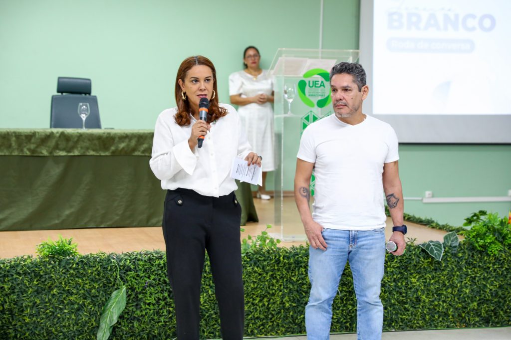 Fotografia de plano médio da vice-reitora da UEA, Prof.ª Dra. Kátia Couceiro, e do palestrante, Prof. Dr. Valber Martins, no palco do Auditório da Reitoria. À esquerda, a vice-reitora segura um microfone e fala para o público; ela veste uma camisa branca e calça preta. À direita, o Prof. Dr. Valber, de camiseta branca e calça de ganga, acompanha a fala. Ao fundo, observa-se parte da tela de projeção com o logótipo da campanha Janeiro Branco e um púlpito de acrílico com o brasão da UEA.