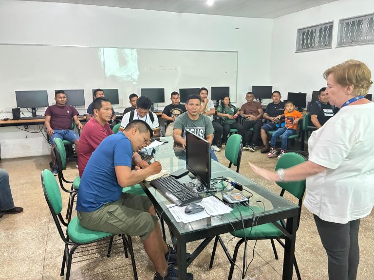 Sala de informática do Centro de Estudos Superiores de São Gabriel da Cachoeira. À direita, em primeiro plano, uma instrutora branca de camiseta branca e cabelos curtos gesticula enquanto fala para um grupo de cerca de 15 professores indígenas das etnias Baniwa e Koripako. À esquerda, um professor de camiseta azul está sentado em uma mesa de vidro, escrevendo atentamente em um caderno. Ao fundo, vários monitores de computador estão alinhados contra a parede branca sob uma tela de projeção.